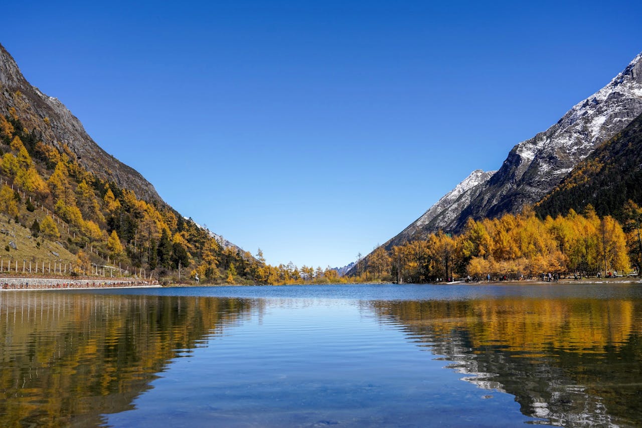 Calm alpine lake with snow-capped mountains and fall foliage reflecting on water.