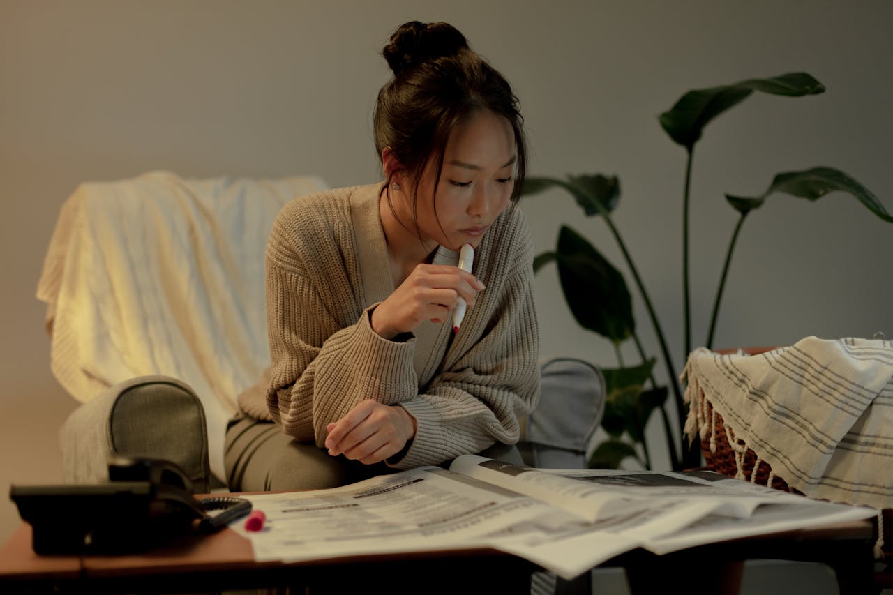 Woman sitting indoors, reading a newspaper, with a thoughtful posture and indoor plants nearby.